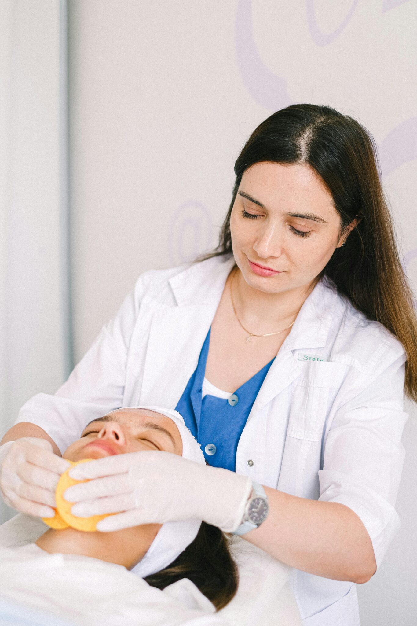Content professional female beauty specialist in white robe cleaning relaxed female customer face with sponged while working in modern light beauty salon